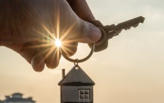 Hand holding key with house keychain backlit by sunset. Homeowners insurance.